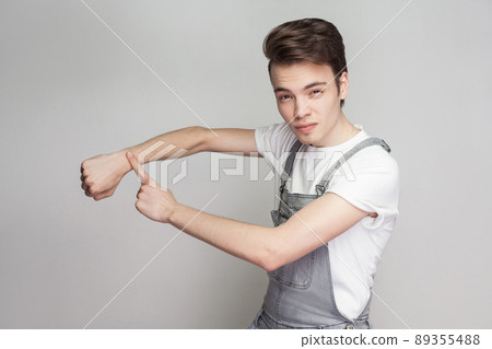 Serious young brunette man in casual style with white t-shirt and denim overalls standing, looking at camera and showing time gesture on hand. indoor studio shot, isolated on gray background. Serious young brunette man in casual style with white t-shirt and denim overalls standing, looking at camera and showing time gesture on hand. indoor studio shot, isolated on gray background. 89355488