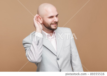 Portrait of cunning handsome middle aged bald bearded businessman in classic light gray suit standing and holding hand on ear and listening. indoor studio shot, isolated on light brown background. Portrait of cunning handsome middle aged bald bearded businessman in classic light gray suit standing and holding hand on ear and listening. indoor studio shot, isolated on light brown background. 89355516