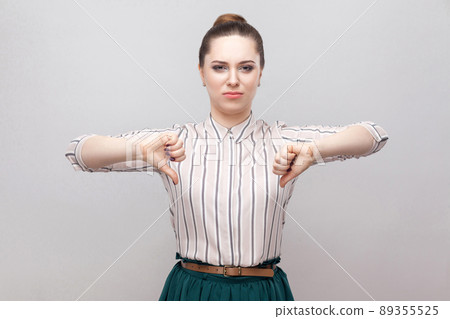 Unhappy beautiful young woman in striped shirt and green skirt with makeup and collected ban hairstyle, standing with thumbs down and looking at camera. indoor studio shot, isolated on grey background Unhappy beautiful young woman in striped shirt and green skirt with makeup and collected ban hairstyle, standing with thumbs down and looking at camera. indoor studio shot, isolated on grey background 89355525