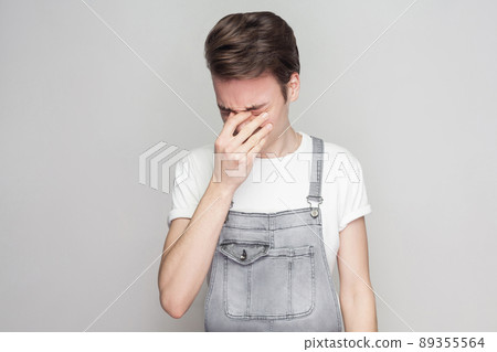 Portrait of unhappy broken heart young brunette man in casual style with white t-shirt and denim overalls standing, holding head down and crying. indoor studio shot, isolated on gray background. Portrait of unhappy broken heart young brunette man in casual style with white t-shirt and denim overalls standing, holding head down and crying. indoor studio shot, isolated on gray background. 89355564