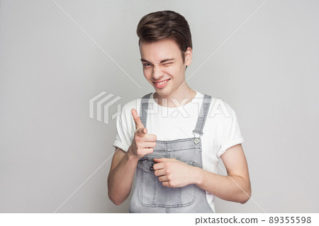 Portrait of funny young brunette man in casual style with t-shirt and denim overalls standing looking and pointing at camera with toothy smile and wink. indoor studio shot, isolated on gray background 89355598