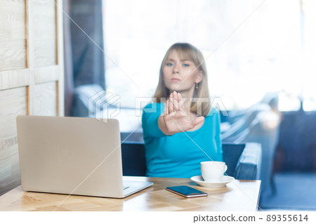 Stop doing it! Portrait of aggressive unhappy young girl freelancer with blonde hair in blue blouse are sitting in cafe and having bad mood and pleased you make pause, showing gesture stop with palm. Stop doing it! Portrait of aggressive unhappy young girl freelancer with blonde hair in blue blouse are sitting in cafe and having bad mood and pleased you make pause, showing gesture stop with palm. 89355614