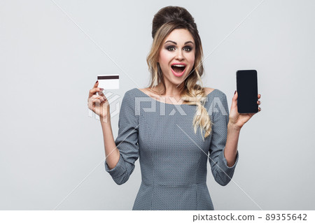 Portrait of happy beautiful modern young girl in grey dress standing, holding cell phone and credit card with open mouth, looking at camera. Indoor, isolated, studio shot, gray background, copy space Portrait of happy beautiful modern young girl in grey dress standing, holding cell phone and credit card with open mouth, looking at camera. Indoor, isolated, studio shot, gray background, copy space 89355642