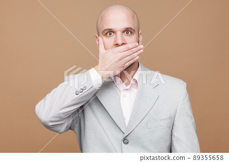 I'm will be quiet. Portrait of shocked handsome middle aged bald bearded businessman in classic light gray suit standing and covered his mouth. indoor studio shot, isolated on light brown background. I'm will be quiet. Portrait of shocked handsome middle aged bald bearded businessman in classic light gray suit standing and covered his mouth. indoor studio shot, isolated on light brown background. 89355658