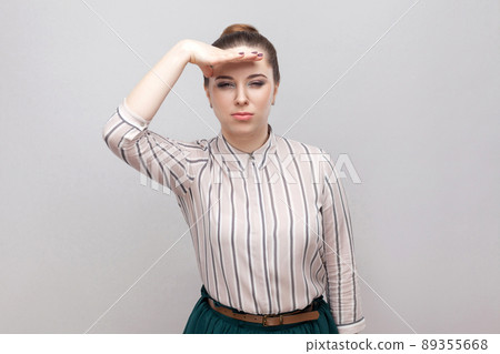 Portrait of beautiful young woman in striped shirt and green skirt with makeup and collected ban hairstyle, standing and holding hand on forehead. indoor studio shot, isolated on grey background. Portrait of beautiful young woman in striped shirt and green skirt with makeup and collected ban hairstyle, standing and holding hand on forehead. indoor studio shot, isolated on grey background. 89355668