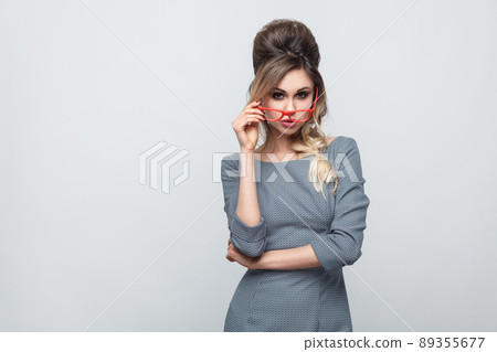 Portrait of dissatisfied beautiful young girl in grey dress with pigtail on head standing, holding red glasses and looking at camera with upset face. Indoor, isolated, studio shot, gray background Portrait of dissatisfied beautiful young girl in grey dress with pigtail on head standing, holding red glasses and looking at camera with upset face. Indoor, isolated, studio shot, gray background 89355677