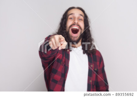 Portrait of amazed handsome man with beard and black long curly hair in casual checkered red shirt standing pointing, looking at camera and surprised. indoor studio shot, isolated on grey background. 89355692