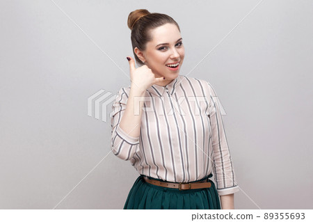 Portrait of funny beautiful young woman in striped shirt with makeup and collected ban hairstyle, standing with call gesture and looking at camera. indoor studio shot, isolated on grey background. Portrait of funny beautiful young woman in striped shirt with makeup and collected ban hairstyle, standing with call gesture and looking at camera. indoor studio shot, isolated on grey background. 89355693