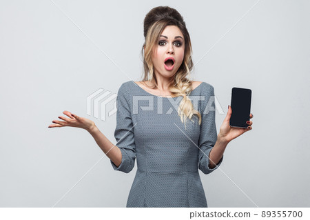 Portrait of shocked beautiful modern young girl in grey dress standing, holding and pointing finger to cell phone and amazed face looking at camera. Indoor, isolated, studio shot, grey background Portrait of shocked beautiful modern young girl in grey dress standing, holding and pointing finger to cell phone and amazed face looking at camera. Indoor, isolated, studio shot, grey background 89355700