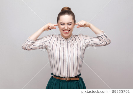 Portrait of unhappy beautiful young woman in striped shirt and green skirt with collected ban hairstyle, standing and putting her finger in ears. indoor studio shot, isolated on grey background. Portrait of unhappy beautiful young woman in striped shirt and green skirt with collected ban hairstyle, standing and putting her finger in ears. indoor studio shot, isolated on grey background. 89355701