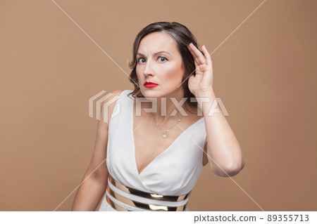 Gossip middle aged woman listen secret. Emotional expressing woman in white dress, red lips and dark curly hairstyle. Studio shot, indoor, isolated on beige or light brown background 89355713