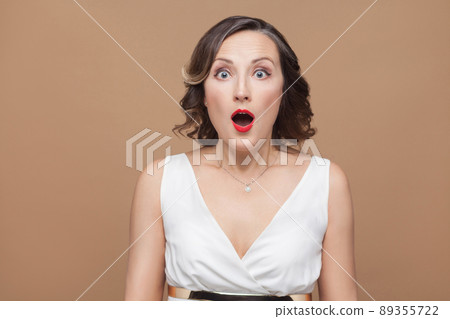 Closeup portrait of shocked middle aged; woman. Emotional expressing woman in white dress, makeup, red lips and dark curly hairstyle. Studio shot, indoor, isolated on beige or light brown background 89355722