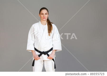 Beautiful athletic young woman in white kimono standing and holding black belt and looking at camera with serious face. Japanese martial arts concept. Indoor, studio shot, isolated on grey background 89355723