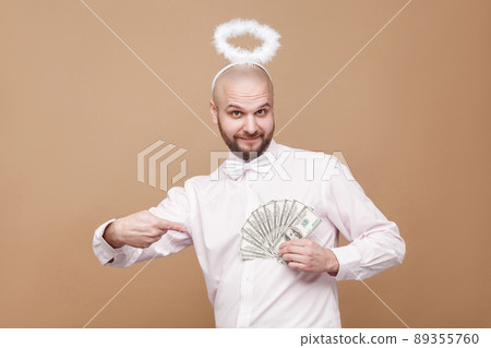 Happy handsome middle aged bald angel in shirt and white halo standing, holding and pointing at many dollars and looking at camera with smile. indoor studio shot, isolated on light brown background. Happy handsome middle aged bald angel in shirt and white halo standing, holding and pointing at many dollars and looking at camera with smile. indoor studio shot, isolated on light brown background. 89355760