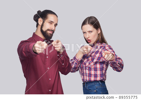 Hey you. Portrait of serious bearded man with black collected hair and woman in casual style standing and pointing at camera with serious face. indoor studio shot, isolated on gray background. Hey you. Portrait of serious bearded man with black collected hair and woman in casual style standing and pointing at camera with serious face. indoor studio shot, isolated on gray background. 89355775