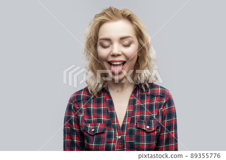 Portrait of funny beautiful blonde young woman in casual red checkered shirt standing with closed eyes and tongue out. indoor studio shot, isolated on grey background. Portrait of funny beautiful blonde young woman in casual red checkered shirt standing with closed eyes and tongue out. indoor studio shot, isolated on grey background. 89355776