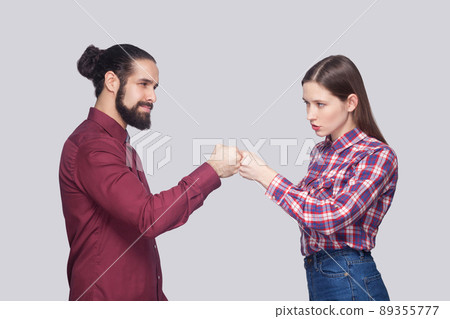 Profile side view of serious bearded man with black collected hair and woman in casual style standing and looking at each other with bump gesture. indoor studio shot, isolated on gray background. Profile side view of serious bearded man with black collected hair and woman in casual style standing and looking at each other with bump gesture. indoor studio shot, isolated on gray background. 89355777