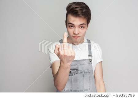 Portrait of crazy angry young brunette man in casual style with white t-shirt and denim overalls looking at camera showing middle finger or fuck sign. indoor studio shot, isolated on gray background. 89355799