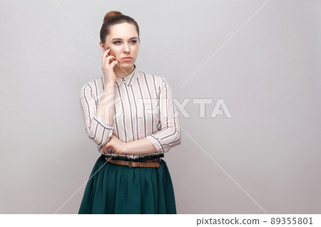 Confused beautiful young woman in striped shirt and green skirt with makeup and collected ban hairstyle, standing thoughtful and looking away. indoor studio shot, isolated on grey background. Confused beautiful young woman in striped shirt and green skirt with makeup and collected ban hairstyle, standing thoughtful and looking away. indoor studio shot, isolated on grey background. 89355801