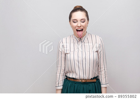 portrait of funny crazy beautiful young woman in striped shirt with makeup and collected ban hairstyle, standing with closed eyes and tongue out. indoor studio shot, isolated on grey background. portrait of funny crazy beautiful young woman in striped shirt with makeup and collected ban hairstyle, standing with closed eyes and tongue out. indoor studio shot, isolated on grey background. 89355802