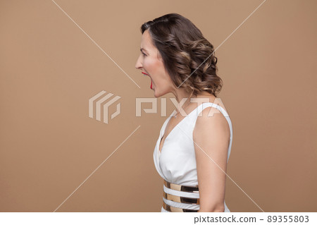 Profile side view of angry middle aged boss woman. Emotional expressing woman in white dress, red lips and dark curly hairstyle. Studio shot, indoor, isolated on beige or light brown background 89355803