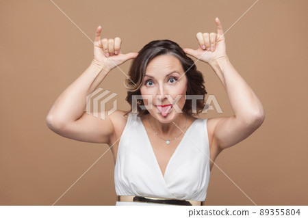 Funny adult woman making stupind mimicry at camera. Emotion and feeling concept. Studio shot, indoor, isolated on light brown background Funny adult woman making stupind mimicry at camera. Emotion and feeling concept. Studio shot, indoor, isolated on light brown background 89355804