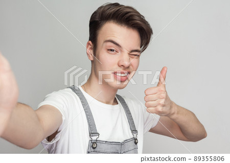 Selfie time! Portrait of happy handsome blogger teenager wearing in demin overalls standing, winking and thump up with toothy smile and making selfie. Indoor, isolated, studio shot, grey background Selfie time! Portrait of happy handsome blogger teenager wearing in demin overalls standing, winking and thump up with toothy smile and making selfie. Indoor, isolated, studio shot, grey background 89355806