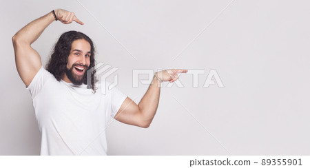 Portrait of strong bodybuilder man in white t-shirt with long wavy hair and beard, standing, looking at camera with toothy smile and pointing at light gray background copy space. indoor studio shot. Portrait of strong bodybuilder man in white t-shirt with long wavy hair and beard, standing, looking at camera with toothy smile and pointing at light gray background copy space. indoor studio shot. 89355901