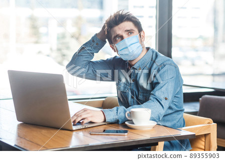 businessman with surgical medical mask in blue shirt sitting and typing on laptop, having new idea and planning own strategy, holding one hand on the head. indoor working and health care concept. 89355903