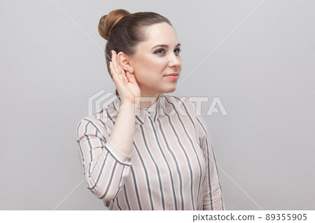 Closeup portrait of funny beautiful young woman in striped shirt and brown collected ban hairstyle, standing with hand near ear and trying to listen. indoor studio shot, isolated on grey background. Closeup portrait of funny beautiful young woman in striped shirt and brown collected ban hairstyle, standing with hand near ear and trying to listen. indoor studio shot, isolated on grey background. 89355905