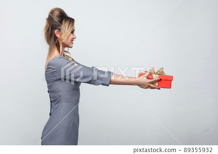 Side view profile of kindness generous beautiful young woman in grey dress standing and giving you present with yellow bow and toothy smile. Indoor, copy space, isolated, studio shot, gray background Side view profile of kindness generous beautiful young woman in grey dress standing and giving you present with yellow bow and toothy smile. Indoor, copy space, isolated, studio shot, gray background 89355932