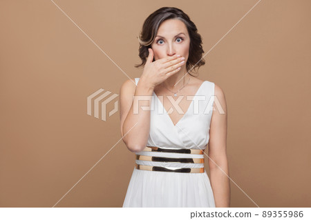 middle aged shocked woman with big eyes closed mouth. Emotional expressing woman in white dress, red lips and dark curly hairstyle. Studio shot, indoor, isolated on beige or light brown background 89355986