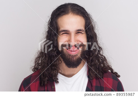 Funny crazy brunette man with beard and black long curly hair in casual style, checkered red shirt standing looking at camera and licking his nose. indoor studio shot, isolated on grey background. 89355987