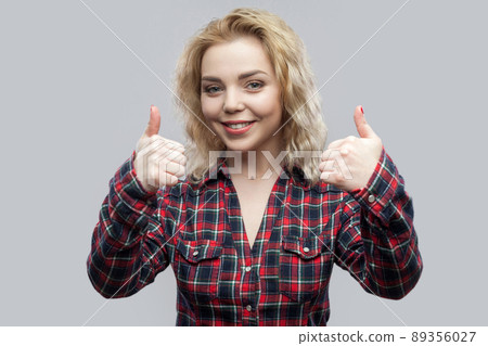 Portrait of happy satisfied beautiful blonde young woman in casual red checkered shirt standing and looking at camera with toothy smile and thumbs up. studio shot, isolated on grey background. 89356027