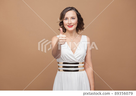 Beautiful adult woman in white dress showing like sign. Emotional expressing woman in white dress, red lips and dark curly hairstyle. Studio shot, indoor, isolated on beige or light brown background Beautiful adult woman in white dress showing like sign. Emotional expressing woman in white dress, red lips and dark curly hairstyle. Studio shot, indoor, isolated on beige or light brown background 89356079