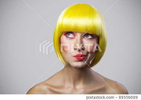 Portrait of pondering cute young woman with freckles, red makeup and yellow wig looking away and thinking about something. indoor studio shot, isolated on gray background. 89356089