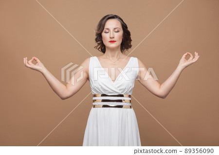 aged woman relaxation, closed eyes and doing asana yoga. Emotional expressing woman in white dress, red lips and dark curly hairstyle. Studio shot, indoor, isolated on beige or light brown background 89356090