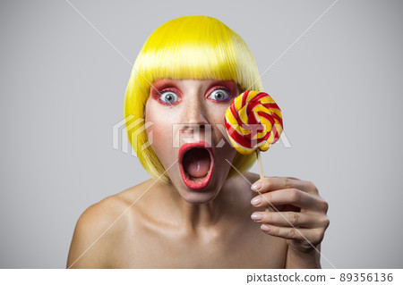 portrait of shocked cute young female model with freckles, red makeup, yellow wig holding colorful candy stick, looking at camera with surprised face. indoor studio shot, isolated on gray background. 89356136