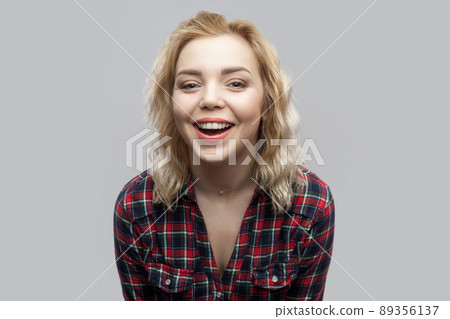 Portrait of happy beautiful blonde young woman in casual red checkered shirt standing, laughing and looking at camera with toothy smile. indoor studio shot, isolated on grey background. 89356137