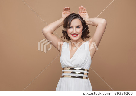 Funny middle aged woman showing rabbit ears and smiling. Emotional expressing woman in white dress, red lips and dark curly hairstyle. Studio shot, indoor, isolated on beige or light brown background 89356206