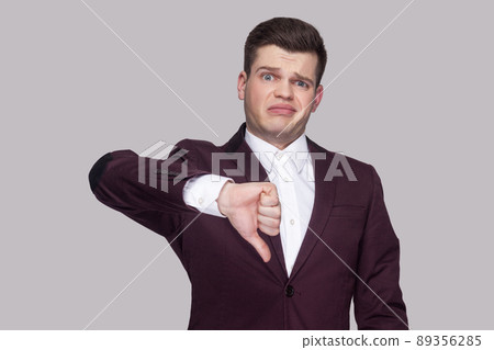 Portrait of handsome confused young man in violet suit and white shirt, standing, looking at camera with thumbs down and unsatisfied face. indoor studio shot, isolated on grey background. 89356285