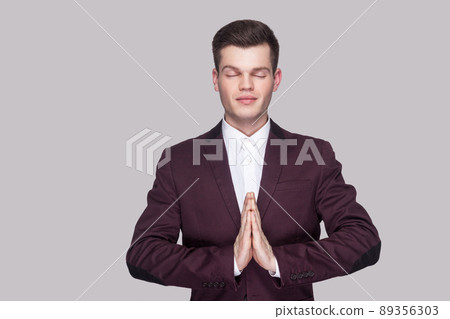 Portrait of calm handsome young man in violet suit and white shirt, standing, closed eyes with yoga gesture and meditation. indoor studio shot, isolated on grey background. 89356303