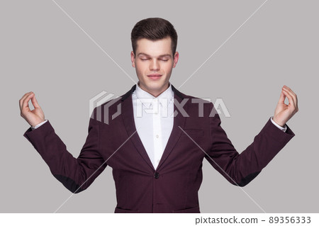 Portrait of calm handsome young man in violet suit and white shirt, standing, raised arms, closed eyes with yoga gesture and meditation. indoor studio shot, isolated on grey background. 89356333