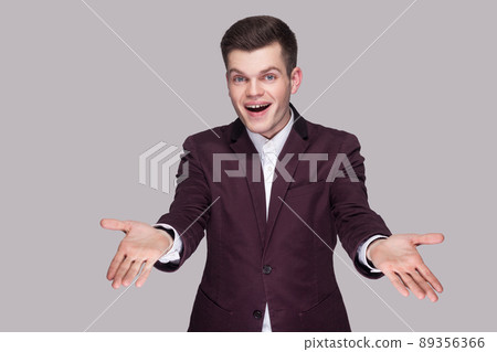 Portrait of happy handsome young man in violet suit and white shirt, standing, looking at camera with raised arms and toothy smile. indoor studio shot, isolated on grey background. 89356366