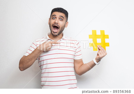 New social media concept. Positive young adult man in t-shirt holding large big yellow hashtag sign and pointing with finger to it with amazing face. Indoor, isolated, studio shot, white background 89356368