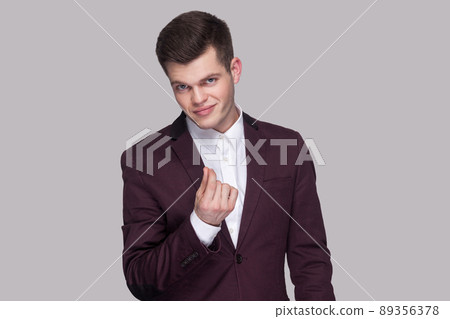 Portrait of serious handsome young man in violet suit and white shirt, standing, looking at camera and showing money gesture. indoor studio shot, isolated on grey background. 89356378