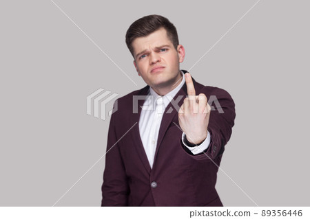 Portrait of angry handsome young man in violet suit and white shirt, standing, looking at camera and showing middle finger. focus on hand. indoor studio shot, isolated on grey background. 89356446