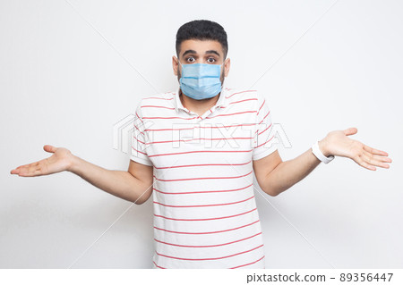 I don't know. Portrait of confused young man with surgical medical mask in striped t-shirt standing with raised arms and dont know what to do. indoor studio shot, isolated on white background. 89356447