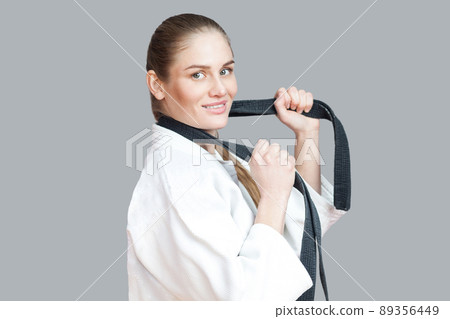 Side view profile of happy beautiful young athletic woman in white kimono with black belt wraps around the neck standing, holding belt and toothy smiling. Indoor studio shot, isolated, grey background 89356449