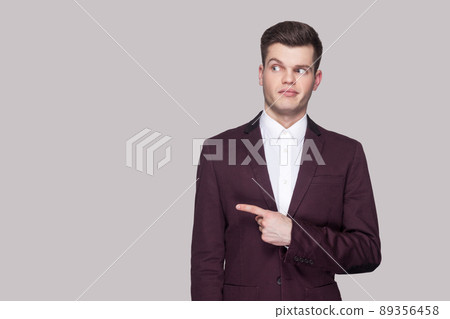 Portrait of funny handsome young man in violet suit and white shirt, standing, pointing and looking away with funny face and smiling. indoor studio shot, isolated on grey background. 89356458